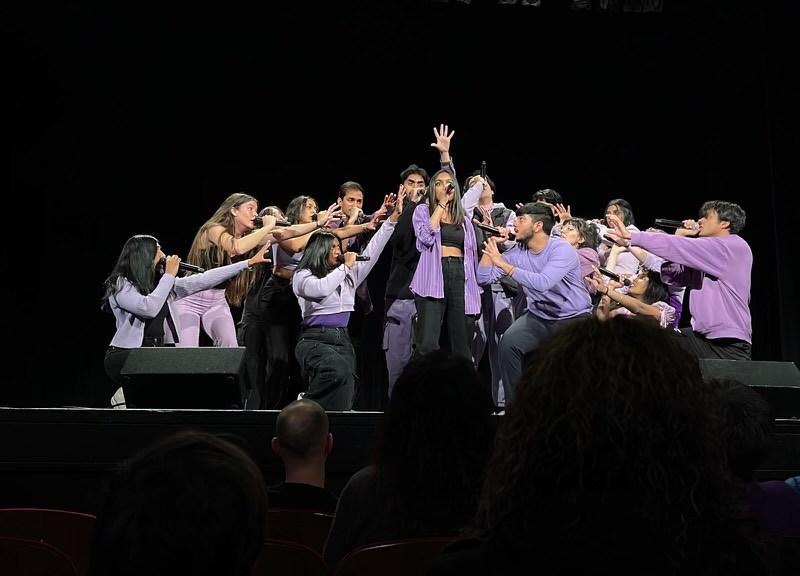 A group of singers come together with mics in their hands on stage. They're singing in matching purple tops, gathering around a group of lead singers.
