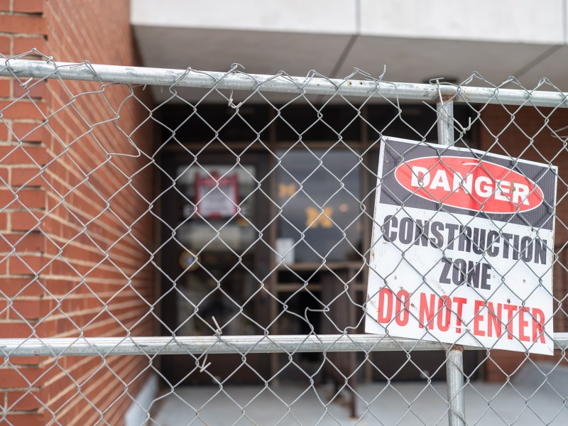 A chain link fence is in focus with a sign saying “Danger. Construction Zone. Do Not Enter”. Behind the fence is the entrance to the CCRB with a yellow block M and a sign warning about asbestos.