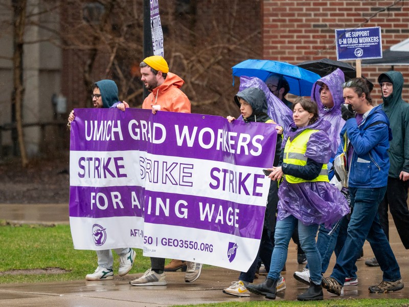 The front of the GEO strike walks bast Hatcher Library with a large purple and white strike banner that says ‘UMich Grad Workers Strike Strike Strike For a Living Wage’.