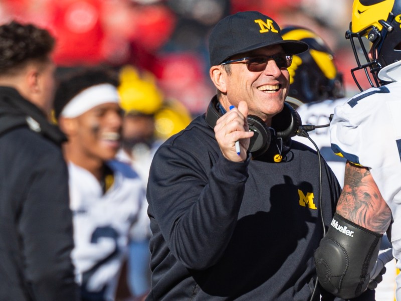 Jim Harbaugh wears a blue jacket and hat with a yellow Block M. He smiles at a Michigan player.