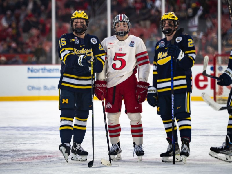 An Ohio State hockey player stands in the middle of two Michigan hockey players on the ice. They hold their sticks.
