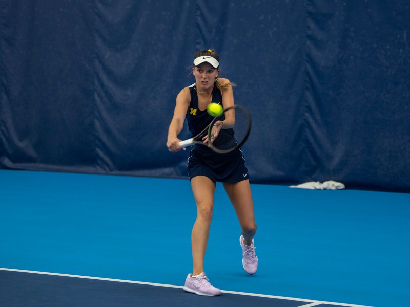 A tennis player holds her racket in a backhand swing to hit a ball to her opponent.