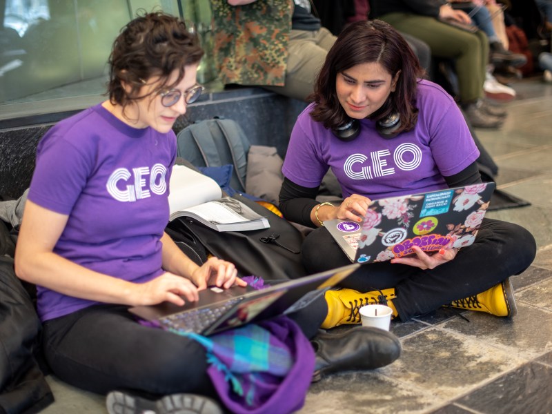 Two women wearing purple GEO shirts sit on the floor of Mason looking at their computers and talking to each other.