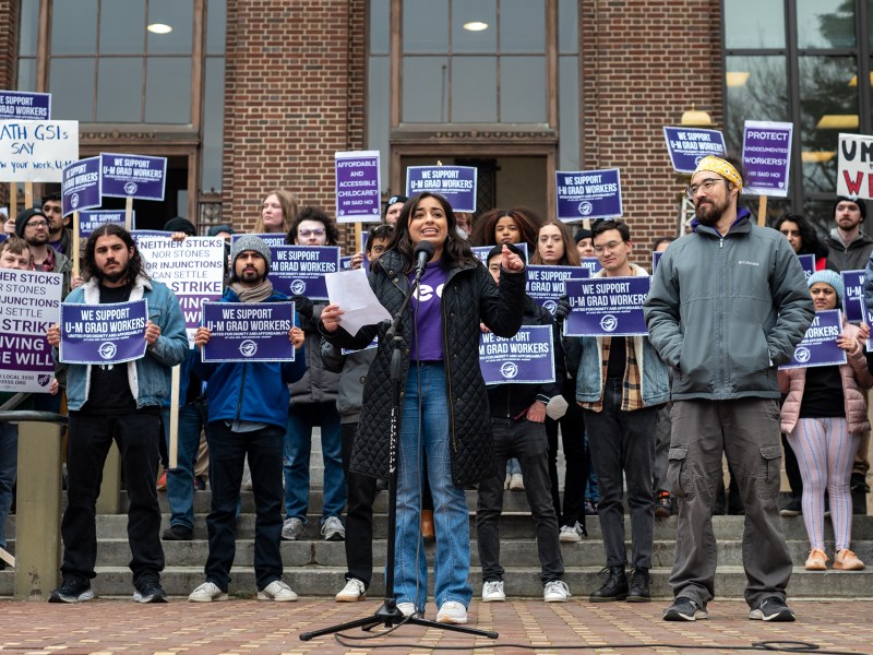 A woman wearing a purple GEO shirt, jeans, and a black winter coat speaks at a microphone on the steps of Hatcher Graduate Library. A crowd of GEO members and allies stand holding purple signs behind her.