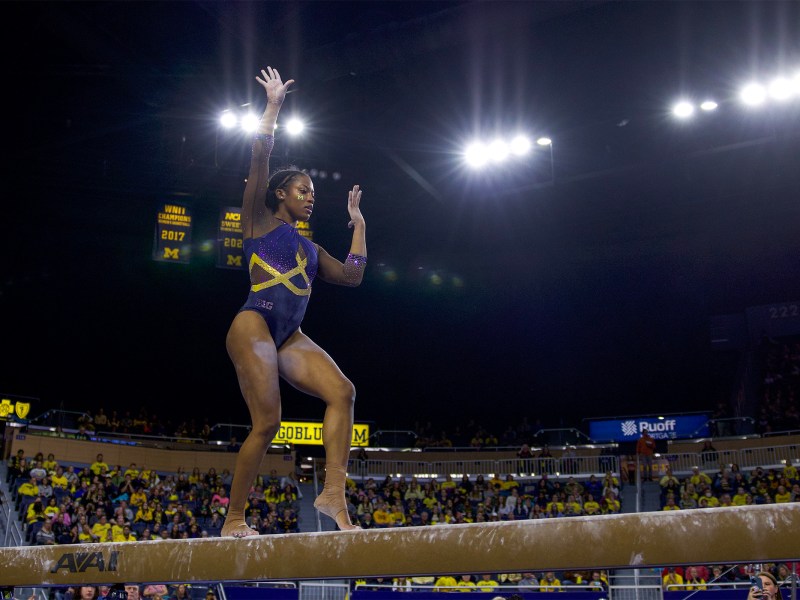 Sierra Brooks poses on the beam with one arm in the air and another bent in front of her face.