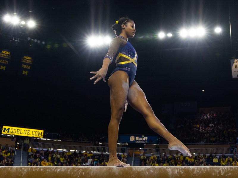 Sierra Brooks is in the middle of a leap on the beam. Her legs are in a split in the air, and her arms are extended outward on both sides. She looks down at the beam to find her landing spot. She wears a purple leotard with a Maize “M” on her cheek.
