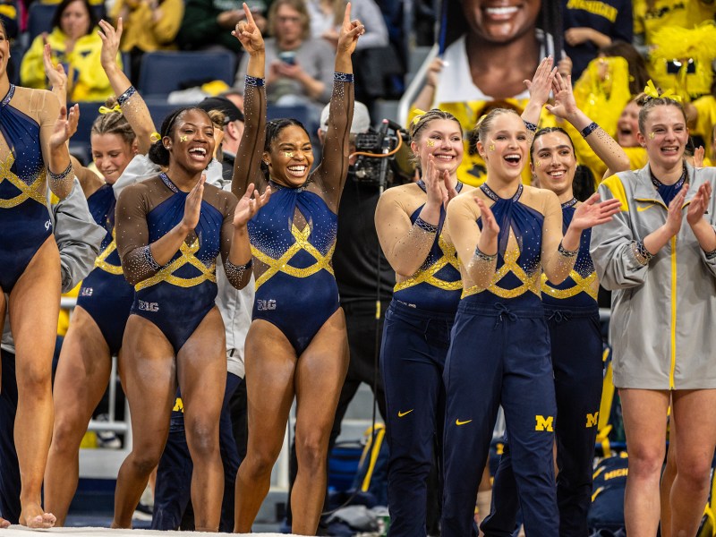 The women’s gymnastics team celebrates on the sideline, with some members clapping and others throwing their hands in the air in excitement. All have smiles on their faces, and wear leotards, though some have sweatpants or sweaters layered on top.