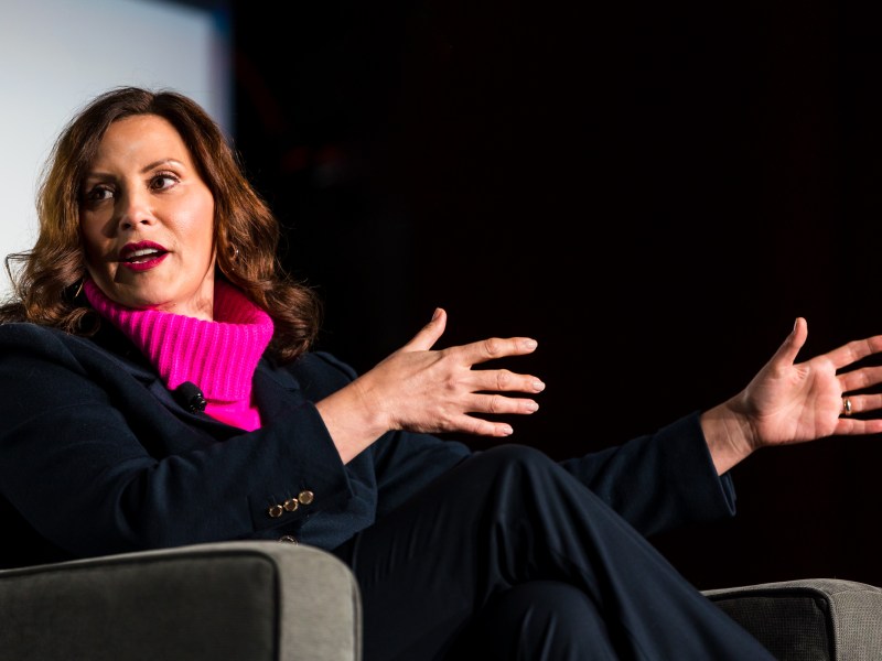 Governor Gretchen Whitmer speaks emphatically with her hands. She is sitting in a chair and looking to her left, wearing a pink turtleneck under a black suit.