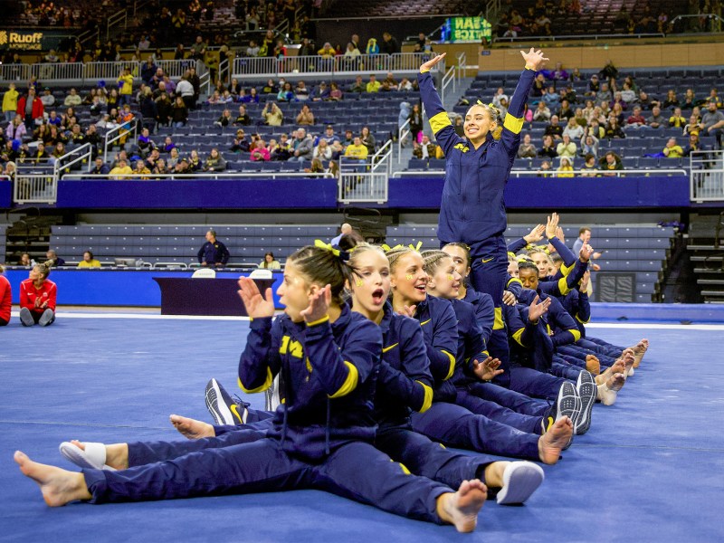 The women's gymnastics team sits in a row on the floor. Natalie Wojcik is standing waving to the crowd.