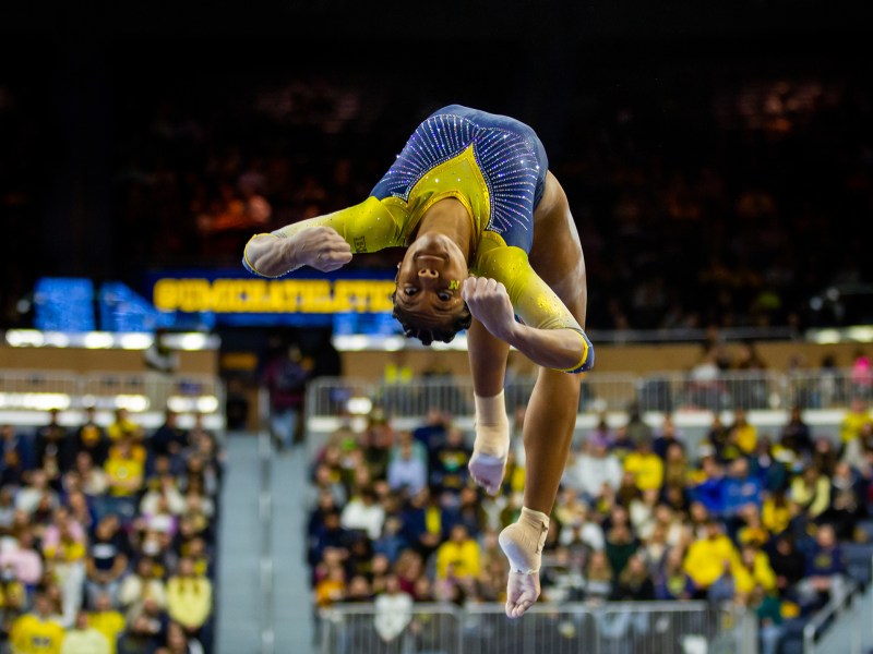 Sierra Brooks flips backwards in a back layout stepout on the beam. She is wearing a maize and blue leotard and behind her are fans sitting in Crisler.