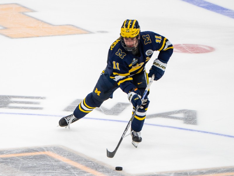 Mackie Samoskevich wears a blue uniform and skates with the puck. The ice has the frozen four logo and block M.