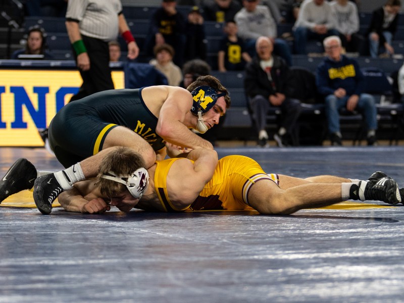 Cole Mattin, a Michigan wrestler, holds down his opponent who is on his stomach. He grabs his opponent's arm and twists it around his back.