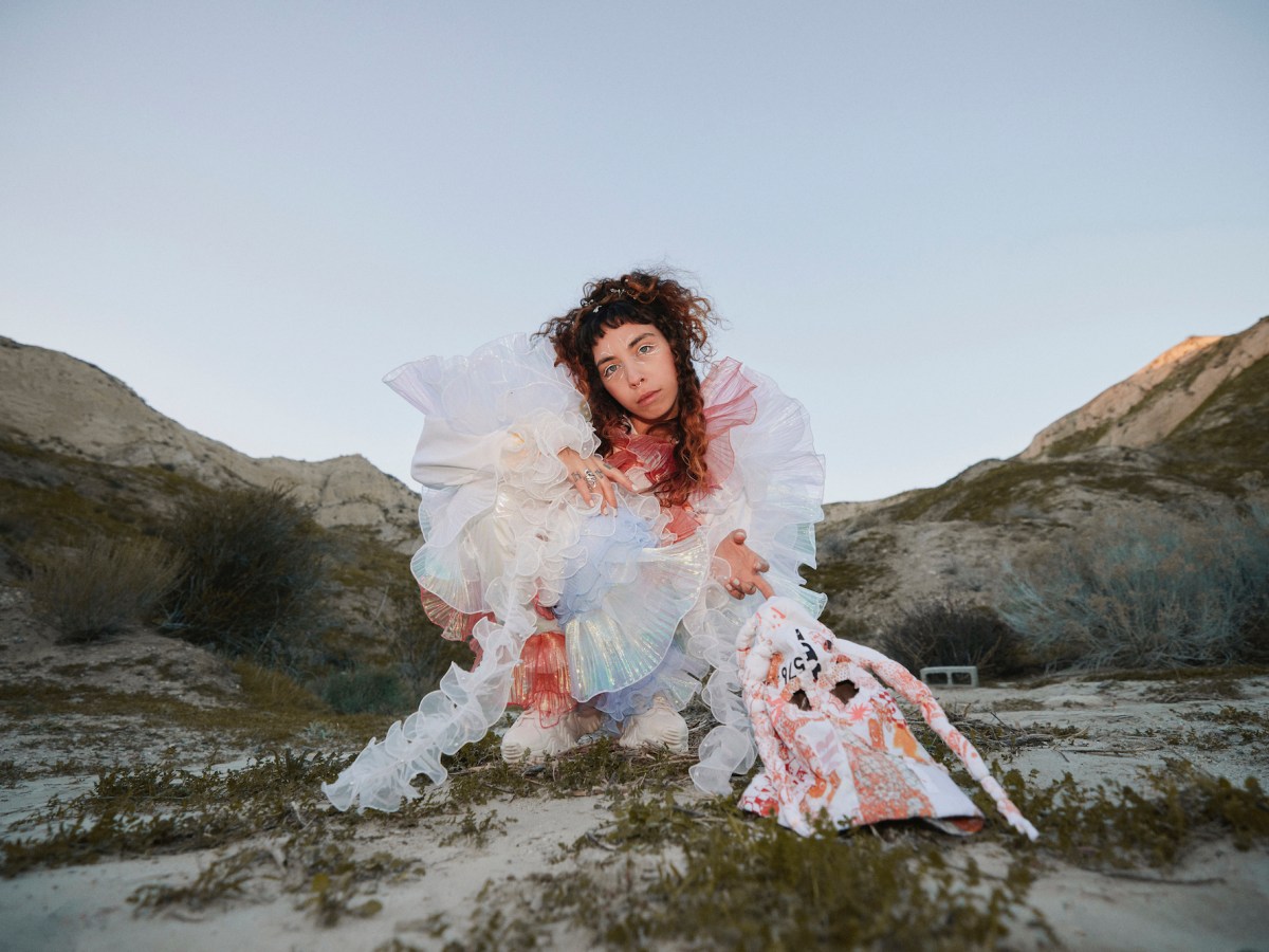 Indigo De Souza poses poses with a white frilly dress with shrub-dotted mountains behind her.