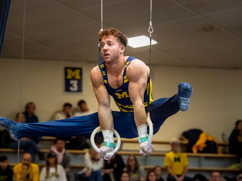 Adam Wooten hangs on the rings with his legs out beside him in the splits.