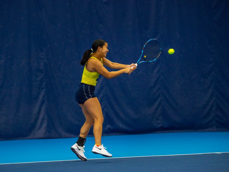 A tennis player reaches her arms across her body while holding her racket up to a ball that’s coming towards her. Her right leg is off the ground and she stands on her left toes.