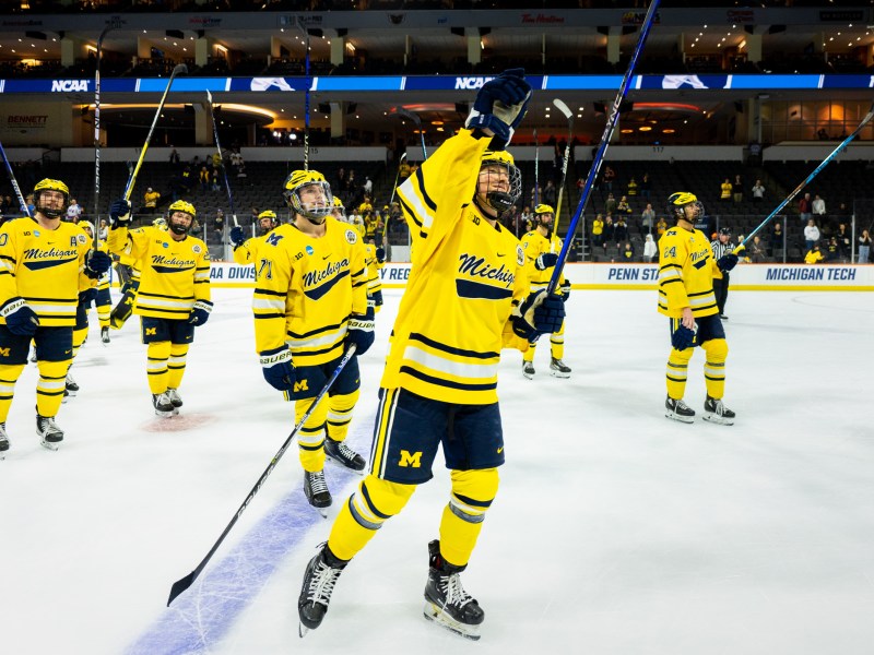 The Michigan Hockey team, wearing yellow jerseys, surrounds the ice in celebration. The player in front of the rest has his right arm raised facing the crowd.
