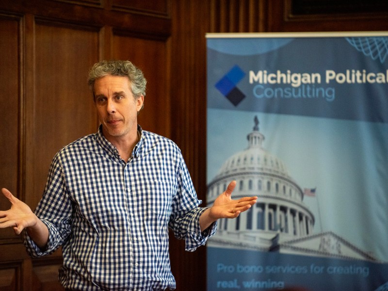 A man holds his hands out and looks to his right as he speaks. A sign for the Michigan Political Consulting event sits to his left.