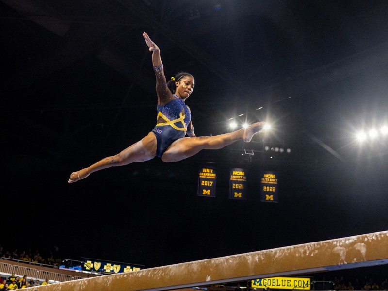 A gymnast jumps into the splits above the balance beam. She looks down at the beam with her arms stretched at her sides and her toes pointed in the direction of both ends of the beam. The lights of Crisler Center shine behind her.