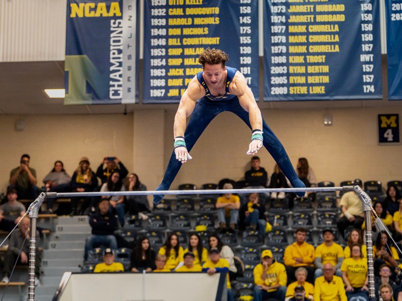 Midair, a Michigan gymnast wearing a blue leotard positions himself to grab the bar as he comes down. Behind him are the NCAA banners and fans sitting in the bleachers.