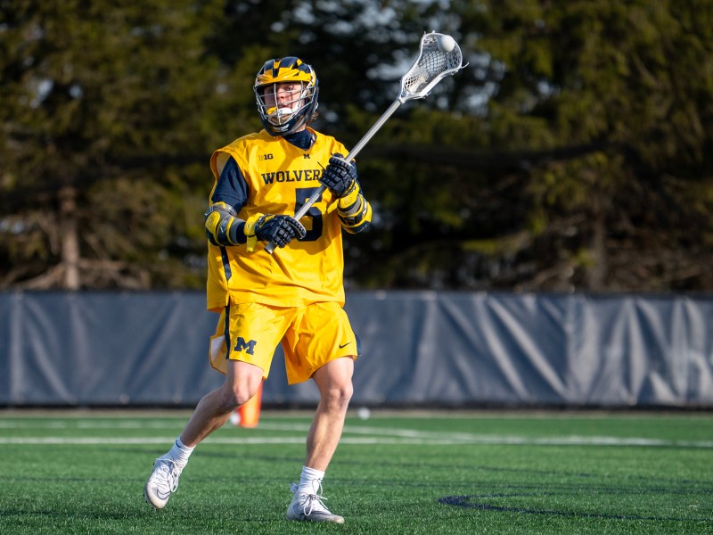 Lacrosse player Michael Boehm wearing a yellow jersey prepares to pass the ball as he moves across the field.