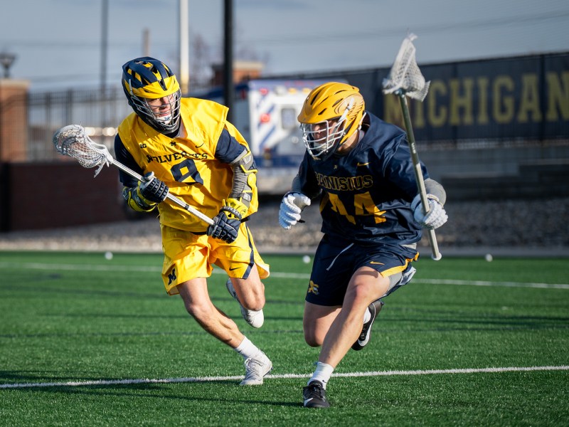 A Michigan men's lacrosse player wearing an all maize uniform runs to guard his opponent wearing all blue.