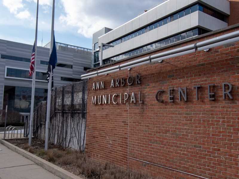 A brick structure stands in front of a glass and metal modern building, with a metal sign labeled “Ann Arbor Municipal Center.” To the left of the sign is a stairway covered in vines, and in front of the stairway is two flagpoles holding the American Flag and the Michigan Flag.