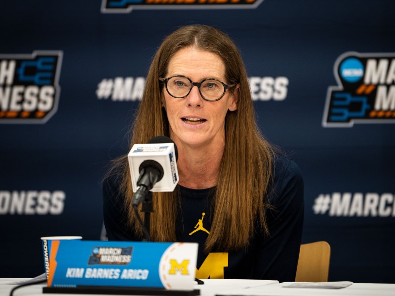 Head Coach Kim Barnes Arico talks into a microphone while taking a seat in front of a media backdrop.