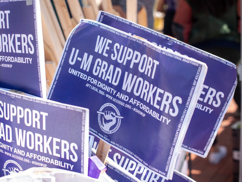 Purple signs saying “We support U-M grad workers” on wooden sticks are in white buckets.