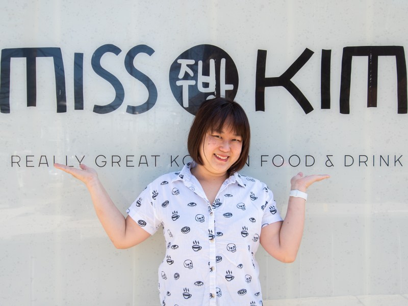 The owner of Miss Kim’s Ji Hye Kim, stands in front of a sign with the name of the restaurant. She is smiling into the camera as she proudly shows off the sign.