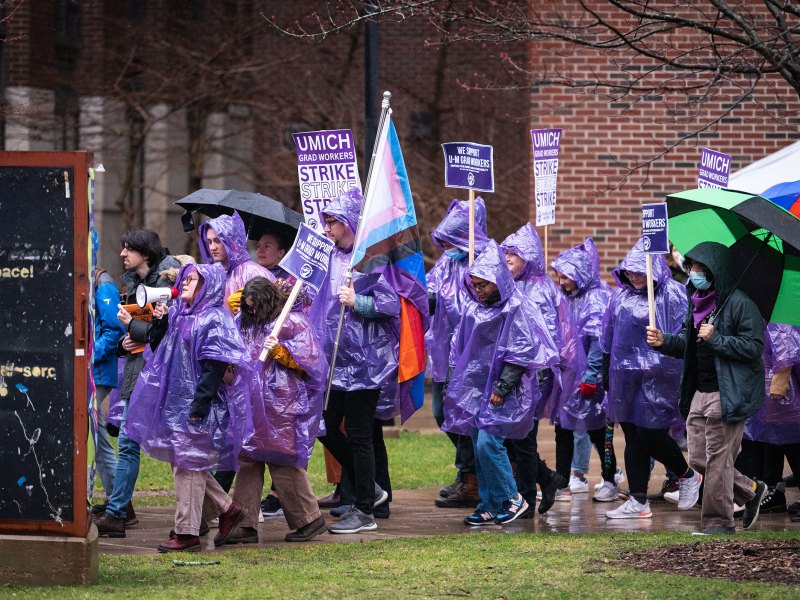 GEO strikers walk down a sidewalk to the left. They are led by a striker holding a megaphone to her mouth. They all wear purple raincoats holding signs.