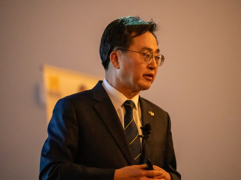 Dong-yeon talk to an audience discussing his life and public service. He is wearing black blazer with a blue tie with thin yellow stripes.