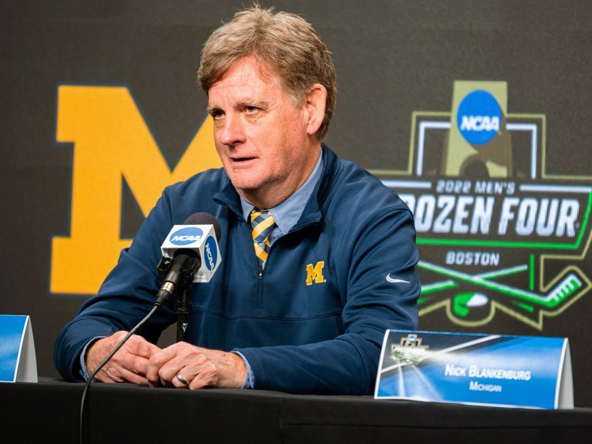 Mel Pearson sits in front of a microphone with the NCAA logo. He is in a blue jacket with a block M and a blue and yellow tie. He is sitting in front of a background promoting the 2022 Men’s Frozen Four for hockey.