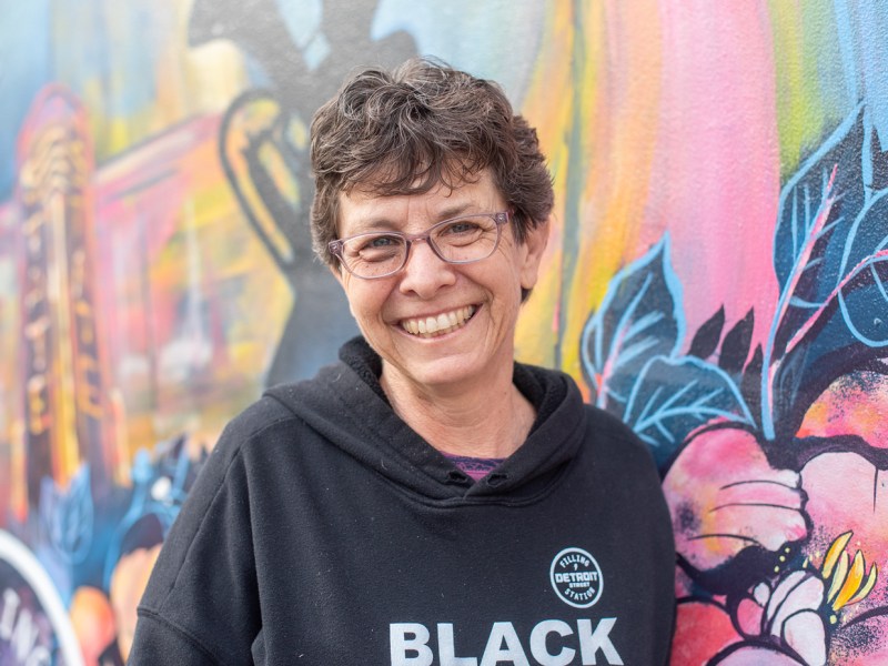 Phillis Engelbert, the owner of Detroit Filling Station, stands in front of a graffiti painted wall wearing a black lives matter sweatshirt. She is smiling into the camera.