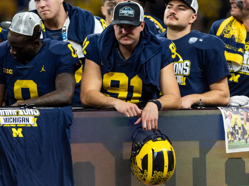 Brad Robbins holds his helmet with his left hand with both arms leaning over an LED sign reading 'Michigan'. His teammates stand beside him and behind him.