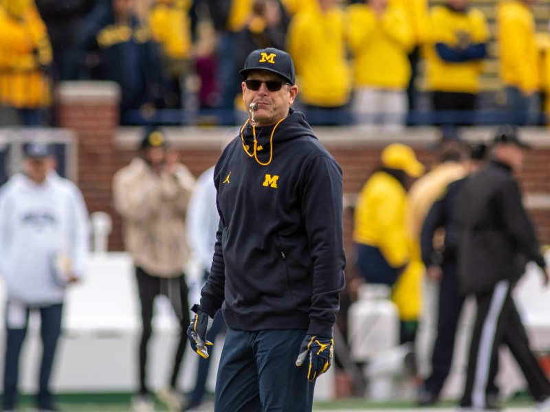 Jim Harbaugh stands on the field with his whistle in his mouth.