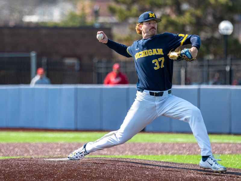 Chase Allen pitches with his right hand behind, along with his right foot behind his left foot. He wears a blue jersey, white pants, and a blue baseball cap with a yellow block M.