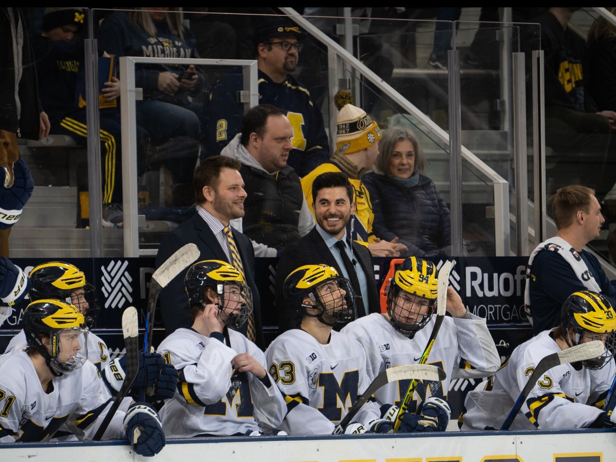 Brandon Naurato and Rob Rassey sit behind the UMich bench. Seven players can be seen sitting in front of them.