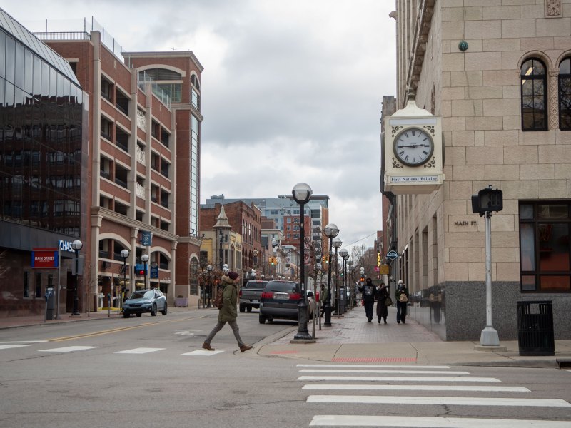 A person crosses the street in downtown Ann Arbor. On a building is a creme clock with a title below that says "First National Building".