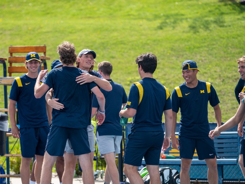 The men's tennis team surrounds each other. Two are embraced in a hug. They wear blue jerseys and blue pants.