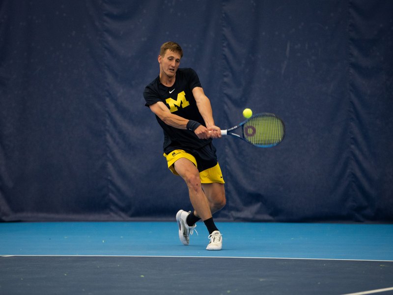 Ondrej Styler aims his racket towards the ball with a backhand with his left leg extended behind him.