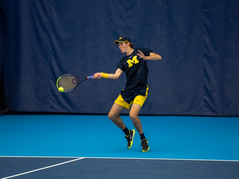 Gavin Young hits the ball with his racket in his right hand. He wears a blue shirt with a yellow block M, a blue hat, and yellow shorts.
