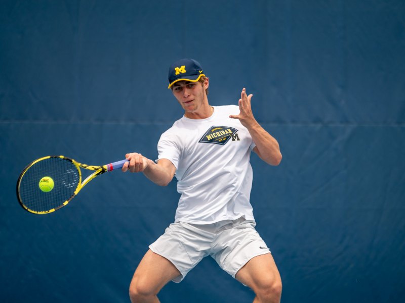 Will Cooksey hits the ball with a racket in his right hand. His left hand is raised. He wears a white shirt and white shorts.