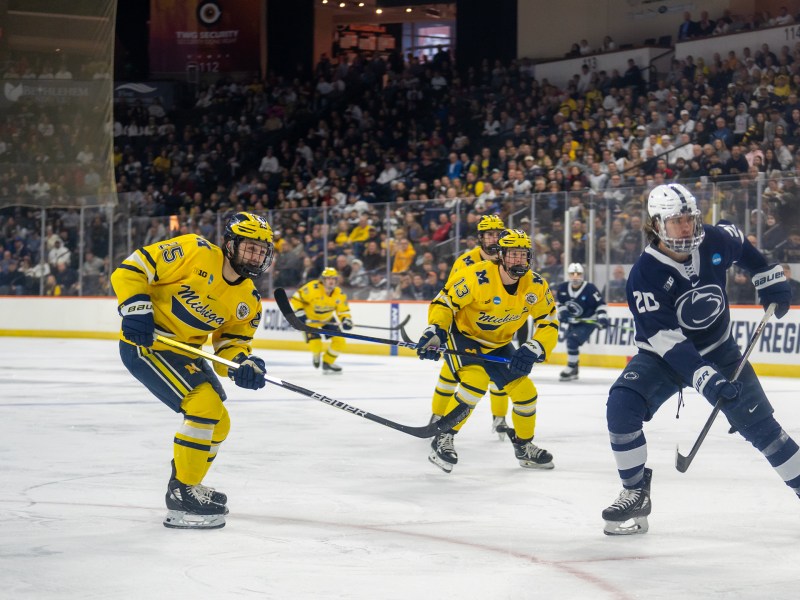 The Michigan hockey team wears yellow jerseys as they skate toward a player on the opposing team dressed in a blue jersey. There are three pictured.