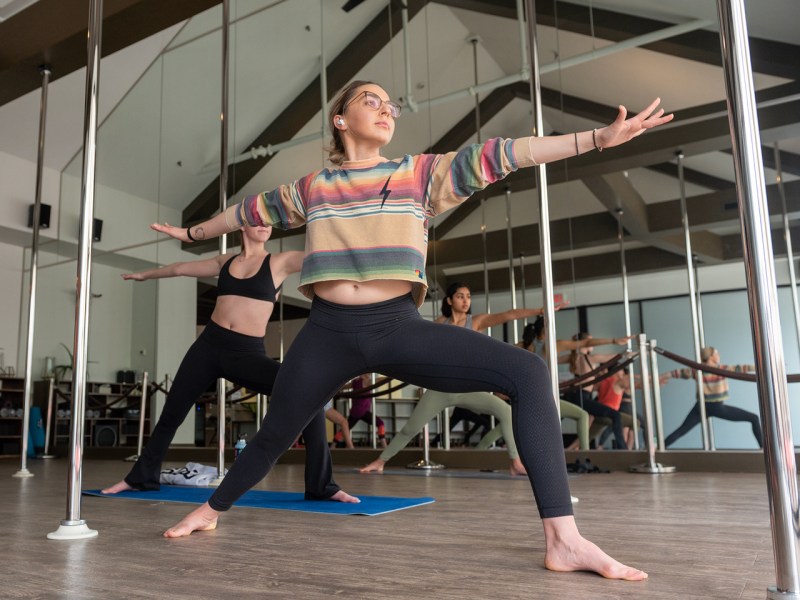 Jessie Lipkowitz, the owner of aUM yoga stands in fierce warrior pose as she leads a yoga class. She is surrounded by mirrors and gray poles.