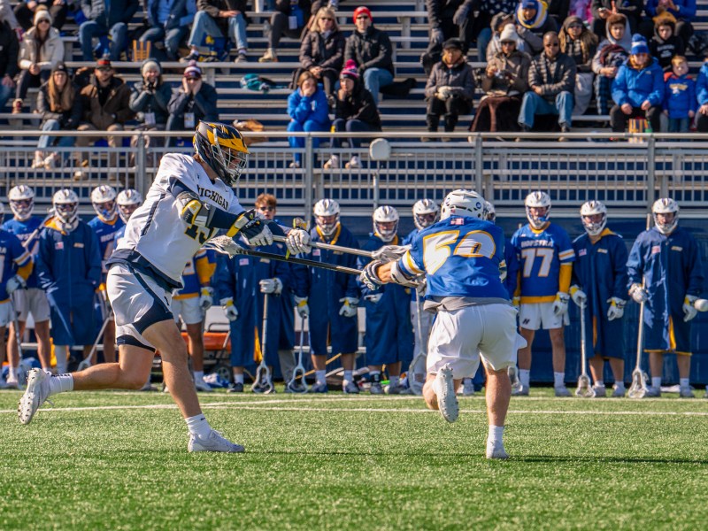 A Michigan men’s lacrosse player in a white uniform bats his lacrosse stick at a Hofstra lacrosse player in a blue uniform. The Hofstra team is standing in the background in front of bleachers where fans are seated.
