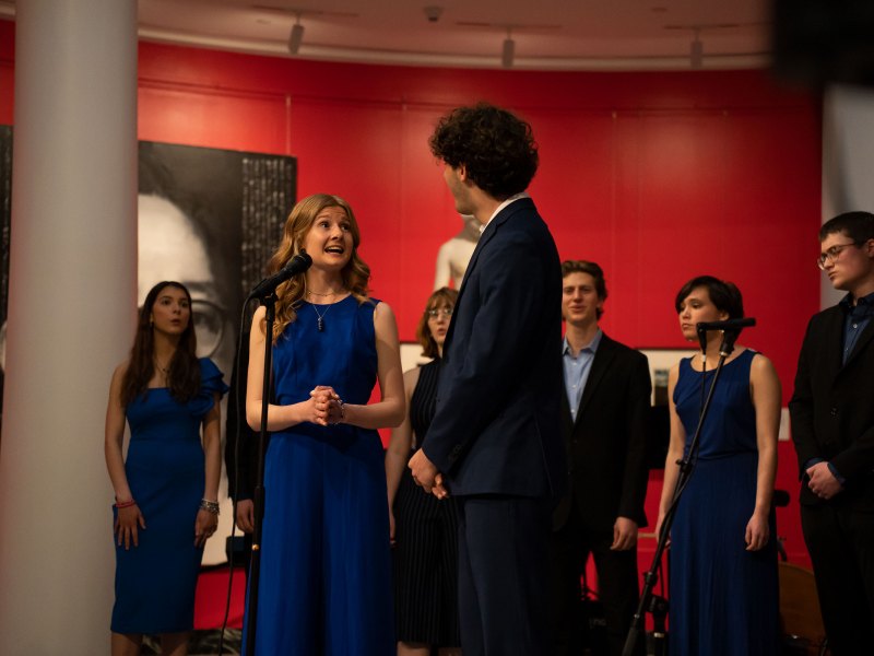 A pair of singers look at each other while performing with the rest of the singers standing in the background.