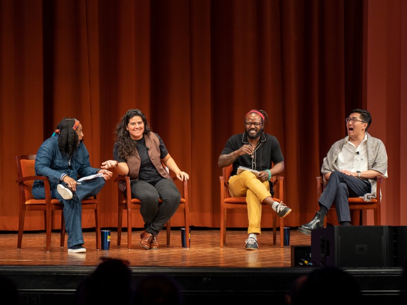 Four speakers sit in a row onstage, each sitting in orange chairs. The speaker on the left wears a denim jumpsuit, the speaker left to center wears a black shirt, a brown vest, and black pants, the speaker to the right center wears yellow pants and a black shirt, and the speaker on the right wears plaid black pants, a white shirt, and a gray cover.