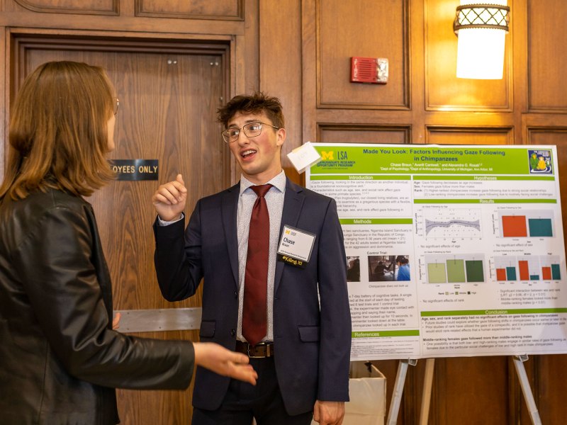 Chase Braun wears a blue suit with a red tie and stands in front of his poster containing information on his research. His right hand is raised as he speaks to a listener.