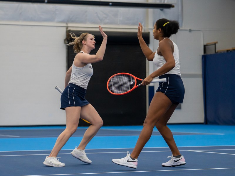 Two michigan womens tennis players go for a high five as they celebrate on the court. They are wearing white tank tops and blue skirts with white shoes.
