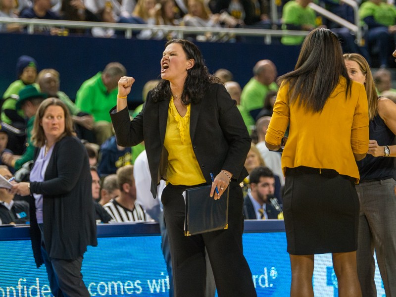 The assistant womens basketball coach screams towards the sideline with her right arm raised in a fist. She is wearing black pants and a black blazer with a yellow top. She is carrying a folder in her left hand.